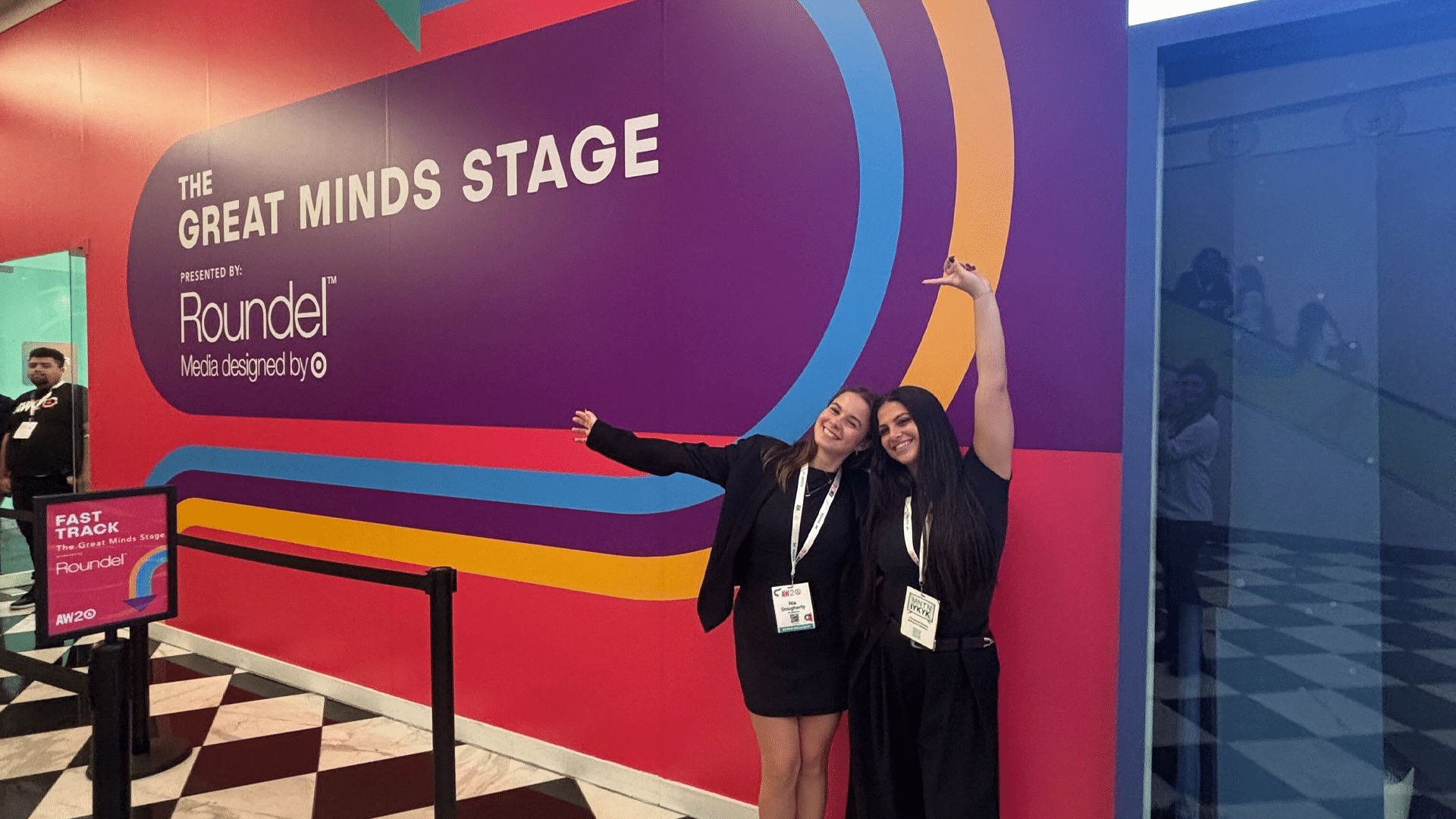 Two women dressed in all black smiling as the point to the Great Minds Stage entrance behind them.