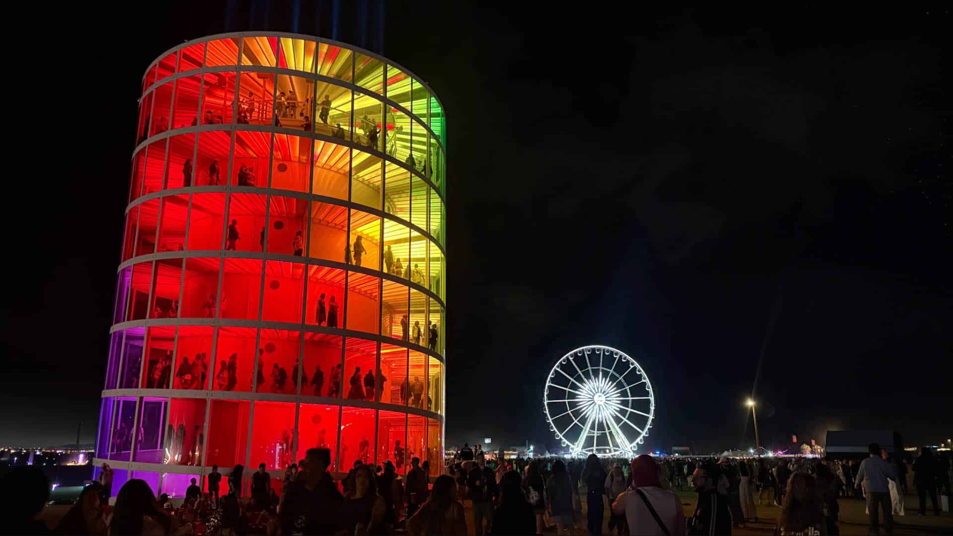 Night view of Spectra's rainbow tower and the illuminated Ferris wheel at Coachella, surrounded by festival crowds under a dark sky.