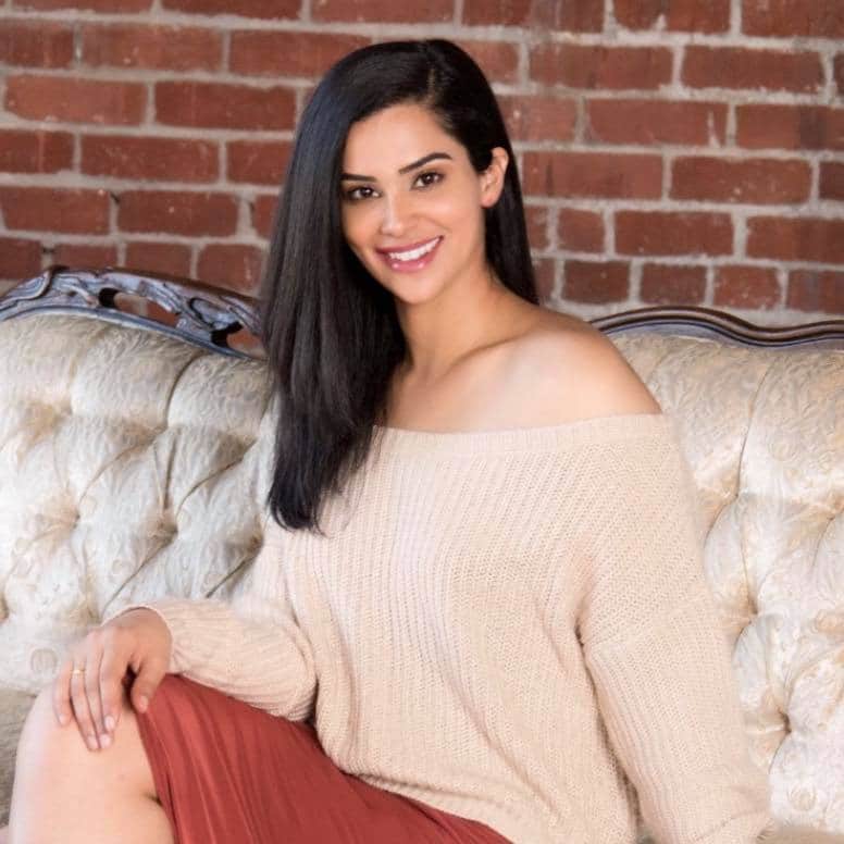 woman with brown hair, sitting on white couch, wearing beige shirt and red skirt smiling